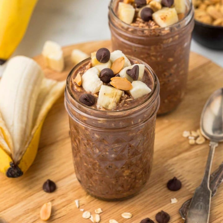 a mason jar with chocolate peanut butter overnight oatmeal topped with banana slices and chocolate chips on a wooden board