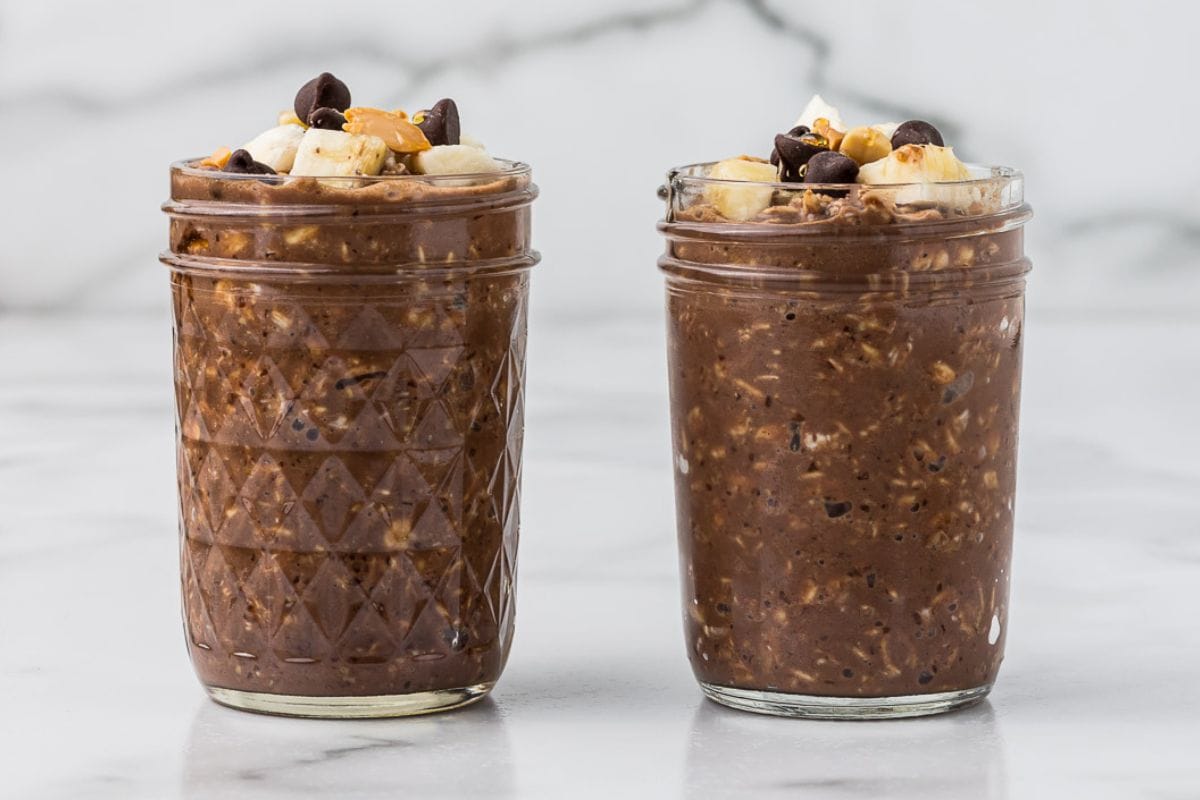 a mason jar with chocolate peanut butter overnight oatmeal topped with banana slices and chocolate chips on a wooden board