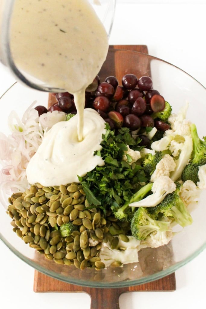 broccoli cauliflower salad ingredients in a large glass bowl with creamy yogurt dressing being poured on top