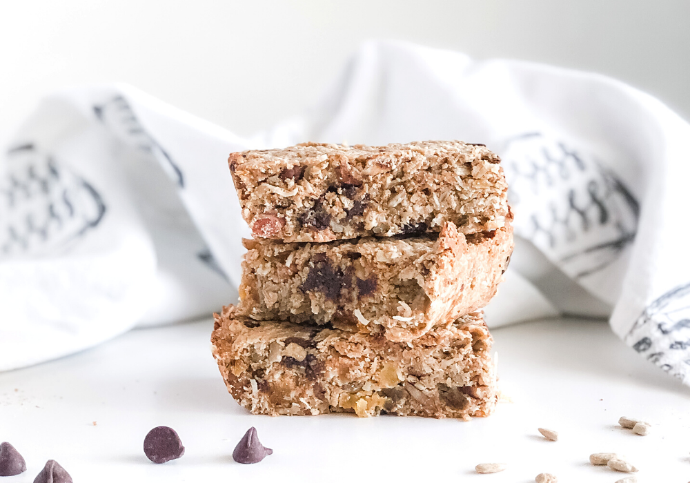 granola bars stacked with chocolate chips and sunflower seeds scattered in front and a kitchen towel behind