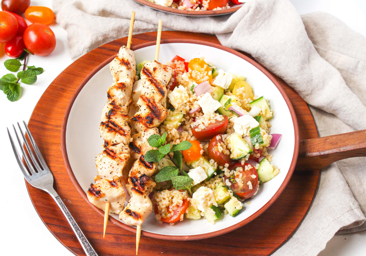 Mediterranean bulgur salad with feta cheese on a white plate next to chicken skewers