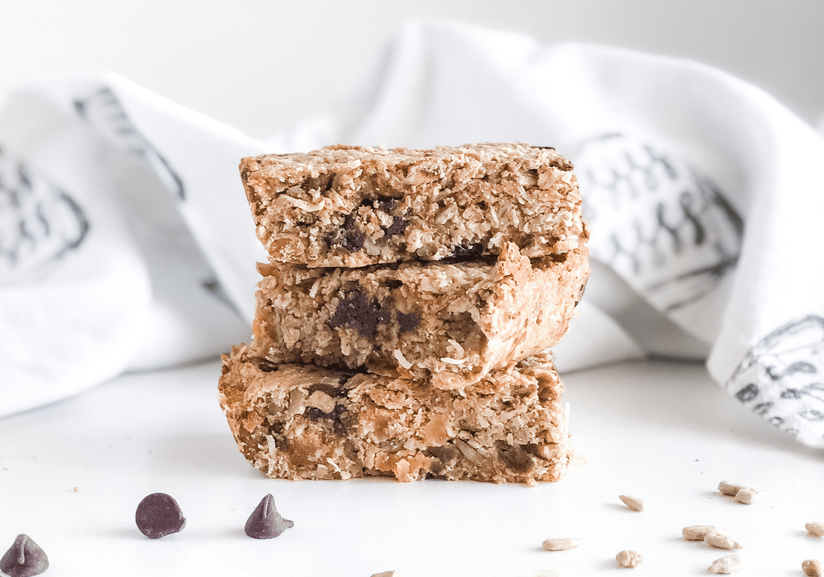 nutty protein granola bars stacked with chocolate chips and sunflower seeds scattered in front and a kitchen towel behind